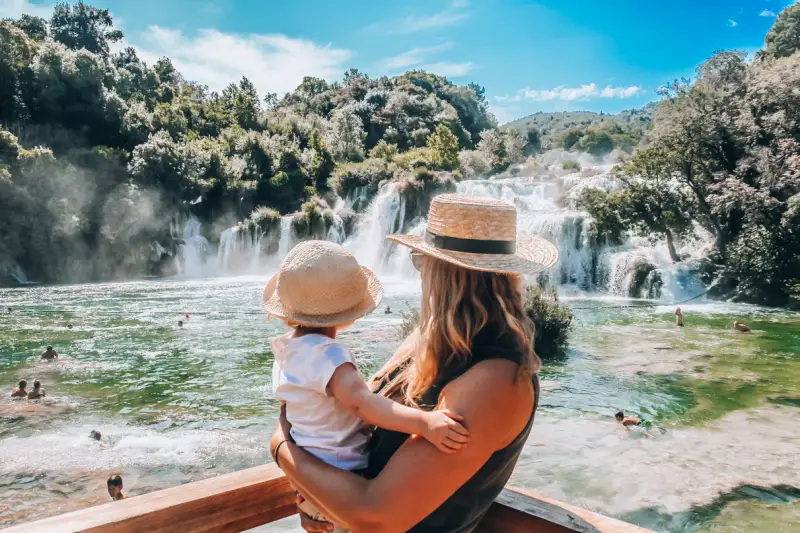 Mama z dzieckiem podziwia wodospady w parku Krka w Chorwacji. Woda jest krystalicznie czysta, a wokół bujna zieleń.