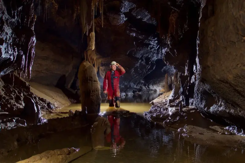 Eksplorator w jaskini, zanurzony po kolana w wodzie, oświetla drogę czołówką. To właśnie jest speleologia – odkrywanie podziemnego świata.